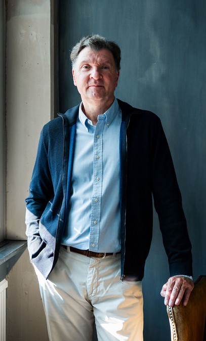 Christian Eibl steht in einer dunklen Strickjacke und hellblauem Hemd vor einer Wand und stützt sich mit einer Hand auf einen Lehnsessel.
